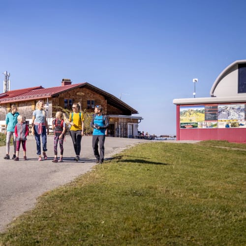 Gruppe von sieben Wanderern spaziert auf einem Weg neben Gebäuden bei sonnigem Wetter.