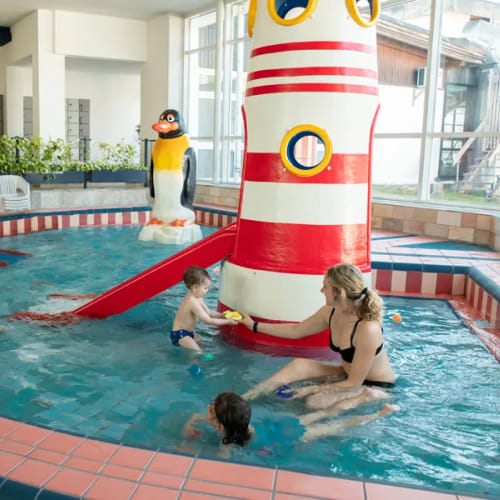 Familie im Hallenbad-Kinderbecken mit roter Rutsche und Pinguin-Dekoration.