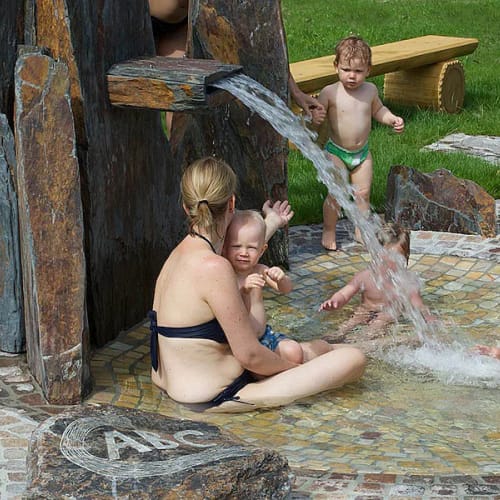 Gruppe Erwachsener und Kleinkinder im Outdoor-Kinderbecken, Wasser aus einer Steinquelle sprudelt.