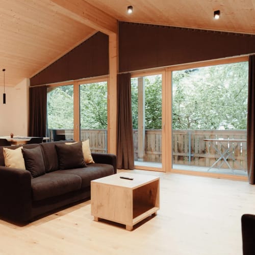 Offenes Wohnzimmer mit Küche, schwarzes Sofa, Holzmöbel und große Fensterfront mit Balkonzugang.