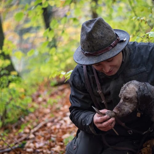 Person mit Hut kniet im Wald und streichelt einen braunen Hund.