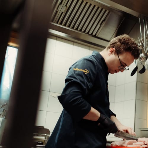 Koch in Arbeitsjacke schneidet Fleisch in einer professionellen Küche.