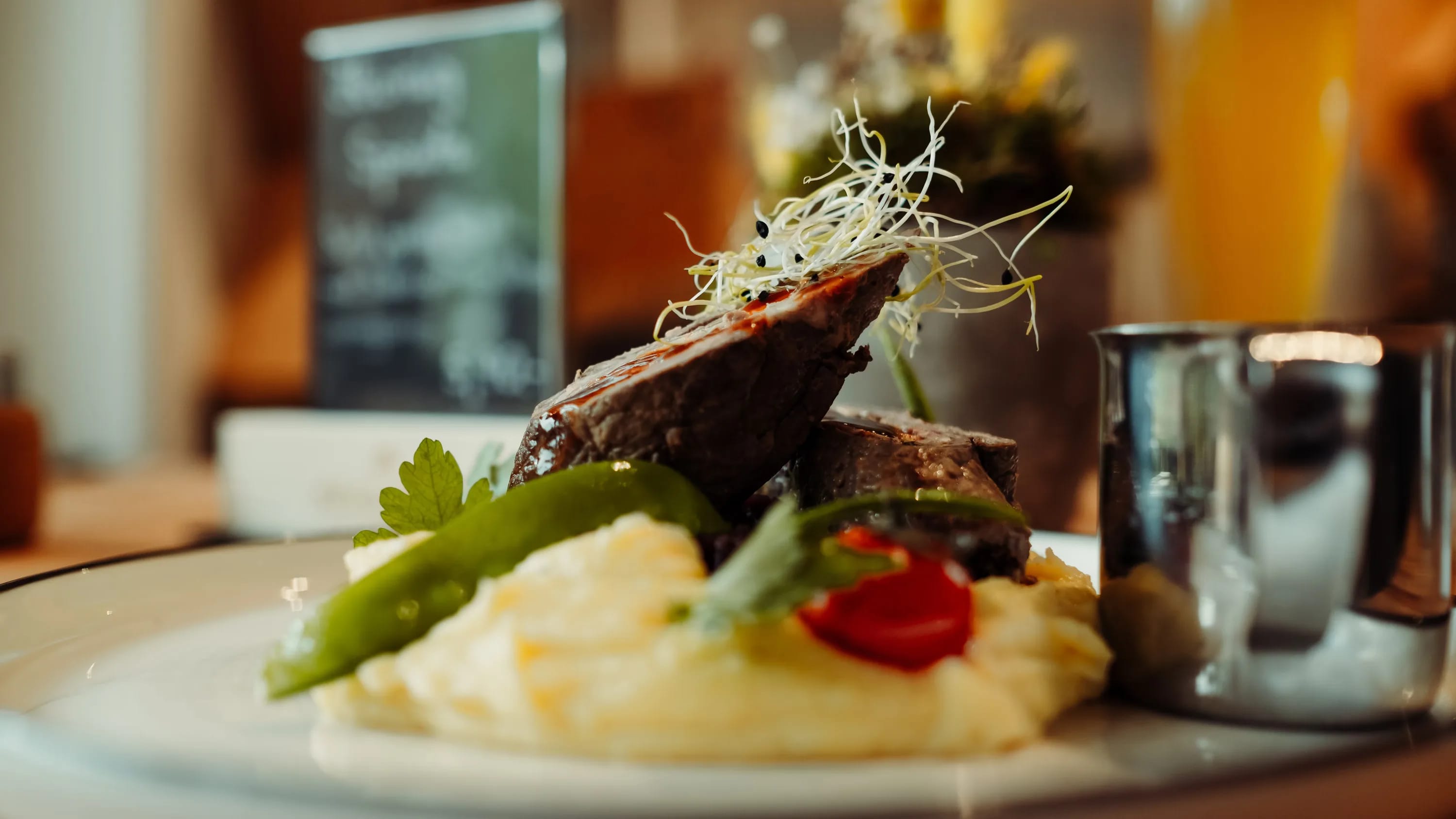 Fleischstück über cremiger Beilage mit grünem Gemüse und Tomate, dekorative Kräuter am Teller