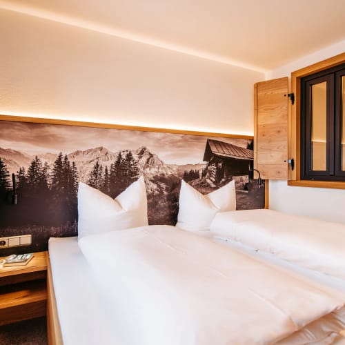 Kleines Schlafzimmer mit zwei Betten, Alpenpanorama-Wandbild, Holzfenster und warmem Licht.