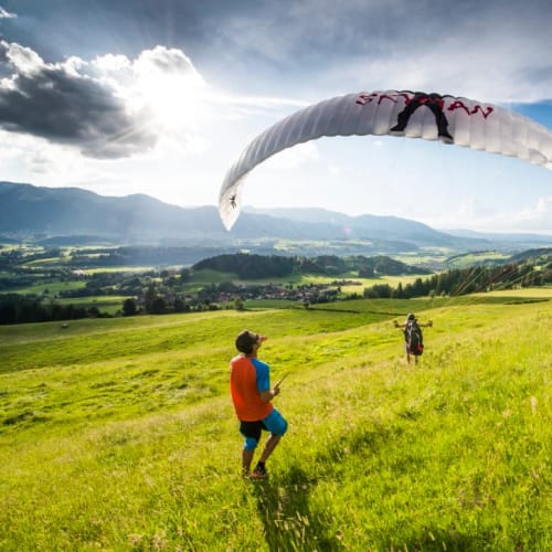 Paragliding-Szene auf grünem Hang; zwei Personen beobachten, Paraglider in der Luft über der Wiese.