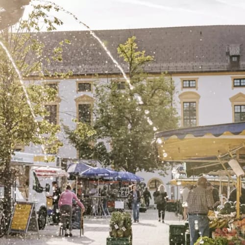 Belebter Marktplatz mit Brunnen, Marktstände und Passanten vor einer historischen Fassade mit Turm.