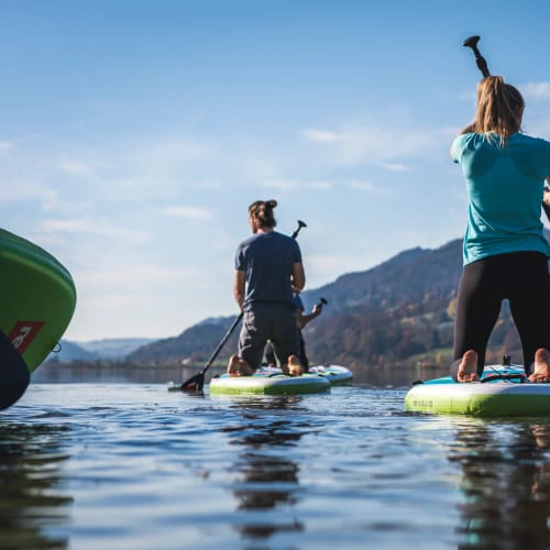 Zwei Personen stehen auf Stand-Up-Paddleboards auf einem ruhigen See, Berge im Hintergrund.