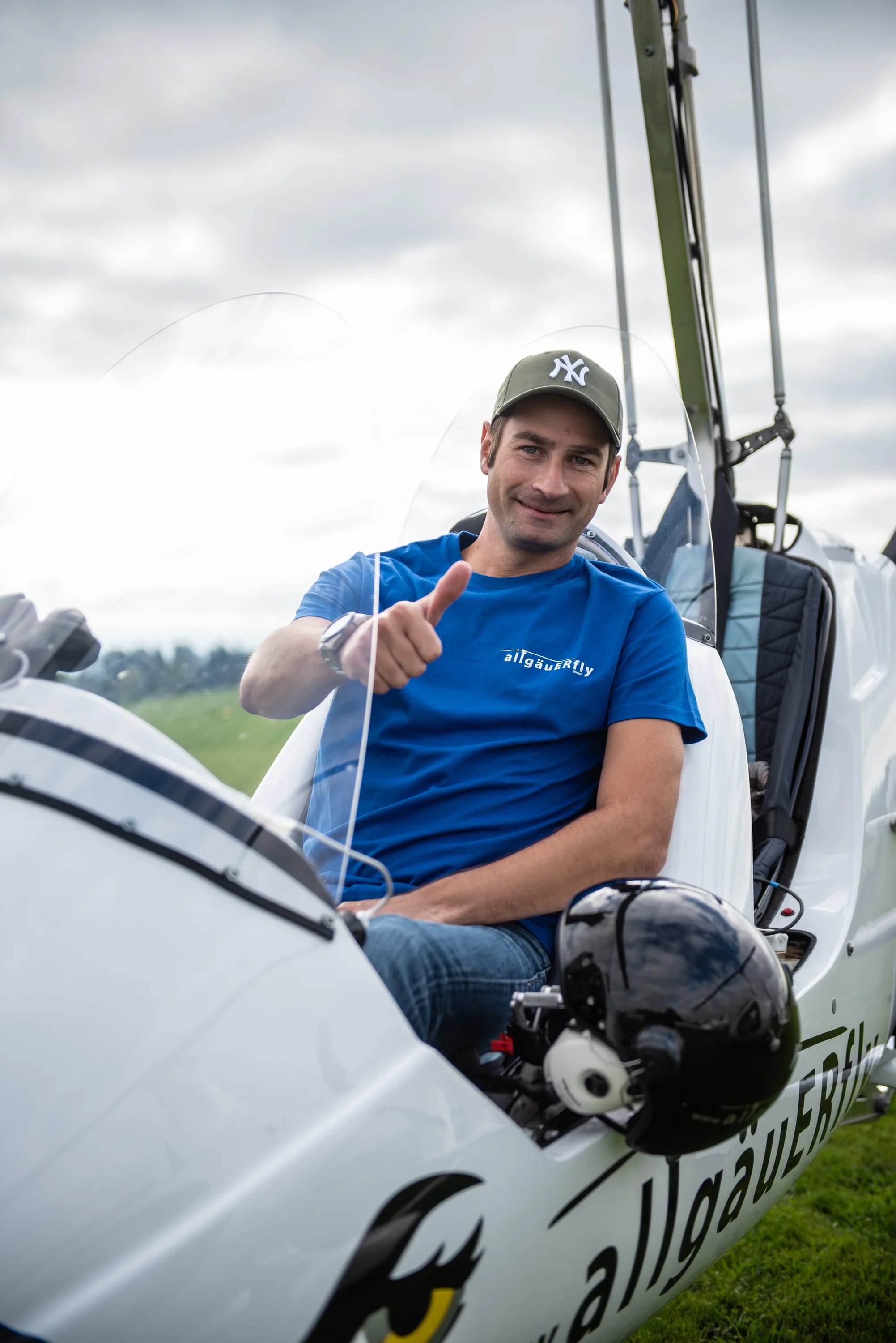 Mann im blauen AllgäuFly-Shirt sitzt in einem Ultraleichtflugzeug und winkt.