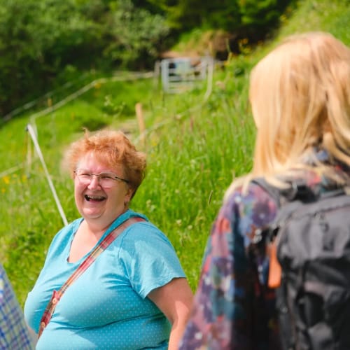 Fröhliche Gruppe im Grünen; zentrale lachende ältere Frau in blauem Shirt im Gespräch mit zwei Bekr