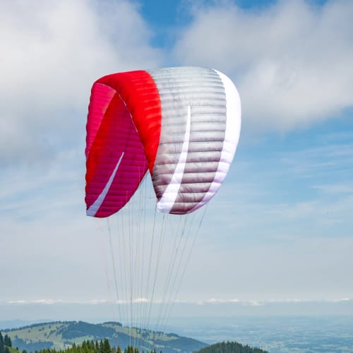Paraglider mit rotem und weißem Schirm startet am Hang über grünem Wald.