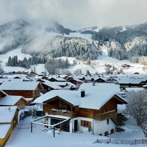 Verschneites Bergdorf mit Holzhäusern und schneebedecktem Wald im Hintergrund.