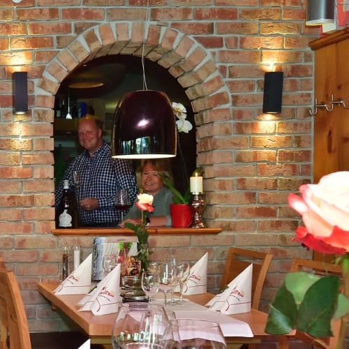 Restaurant mit Backsteinwand und drei Bogenfenstern, Personen hinter Fenstern, gedeckte Tische.