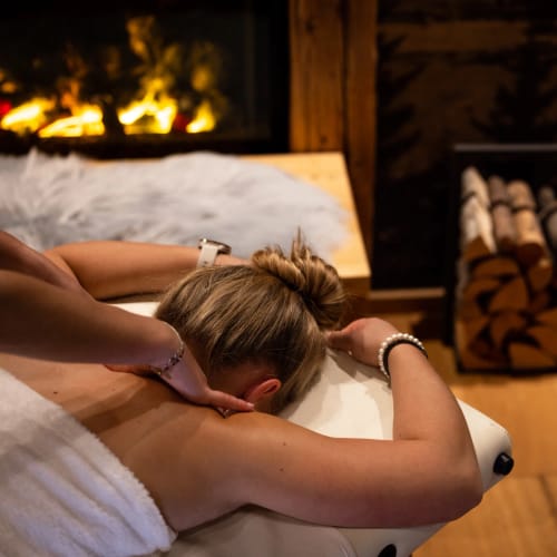 Person liegt entspannt auf Massagebank vor Kamin; Holzstapel im Hintergrund.