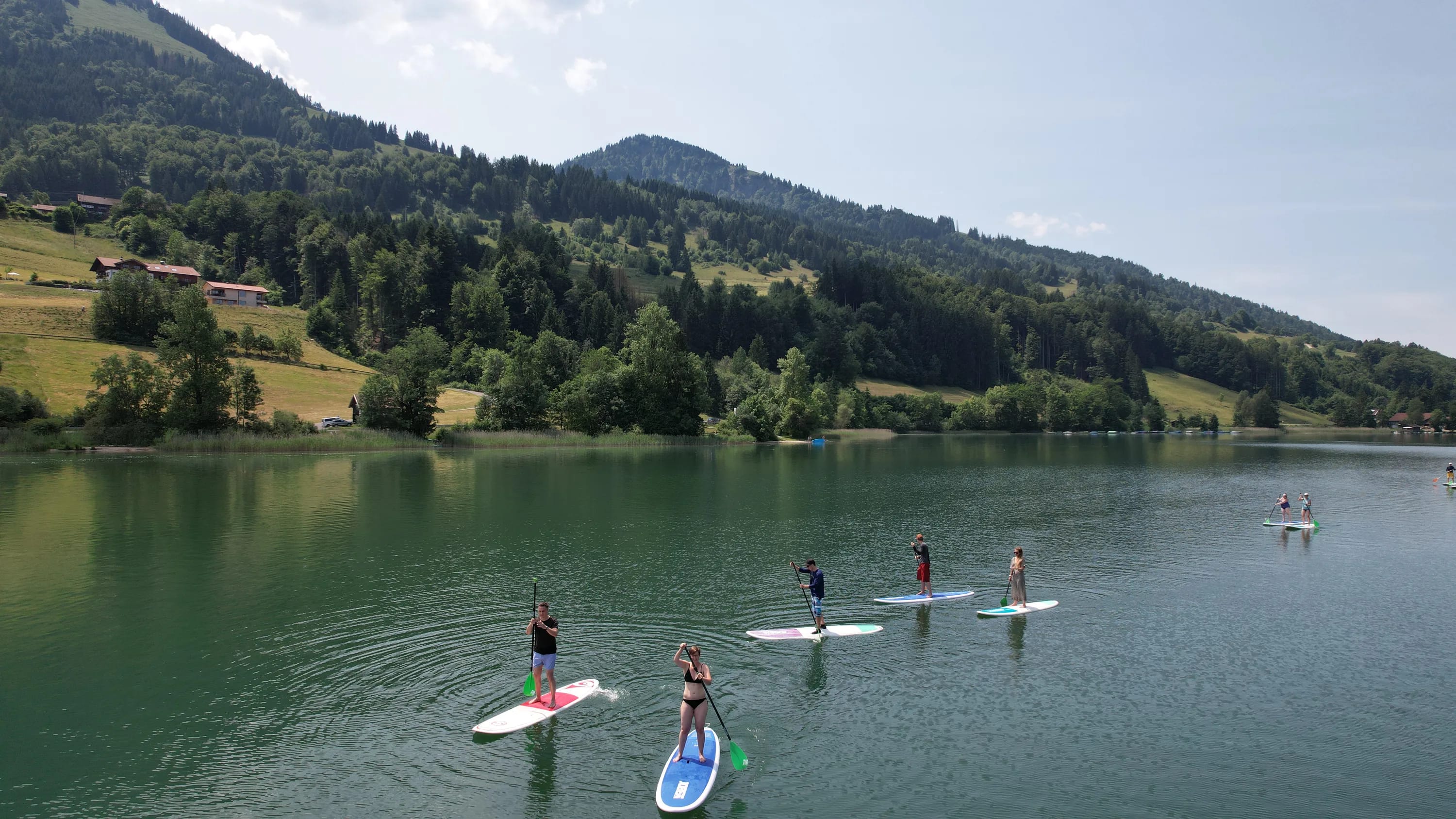 Mehrere Stand-Up-Paddler auf einem ruhigen See, umgeben von grünen Hügeln und Wald.