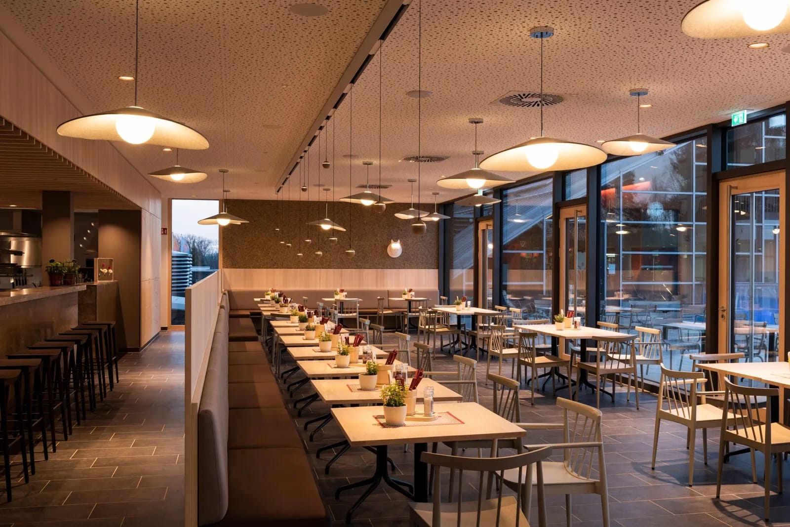 Heller Restaurantbereich mit Reihen von Tischen und Stühlen, gemütlicher Beleuchtung und großen Fen.