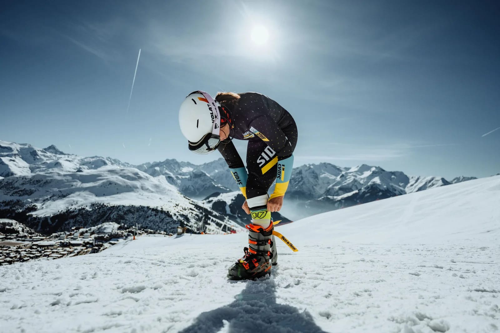 Skifahrer in Helm kniet auf Schnee und justiert Skibindung, Alpen im Hintergrund.