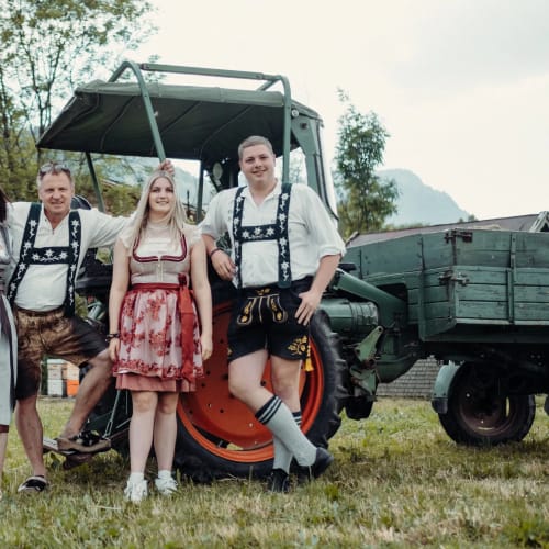 Vier Personen in Trachten vor Traktor und Anhänger auf einer Wiese.