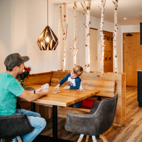 Familie beim Spielen am Esstisch in einer alpinen Stube, Mutter kocht in der Küche.