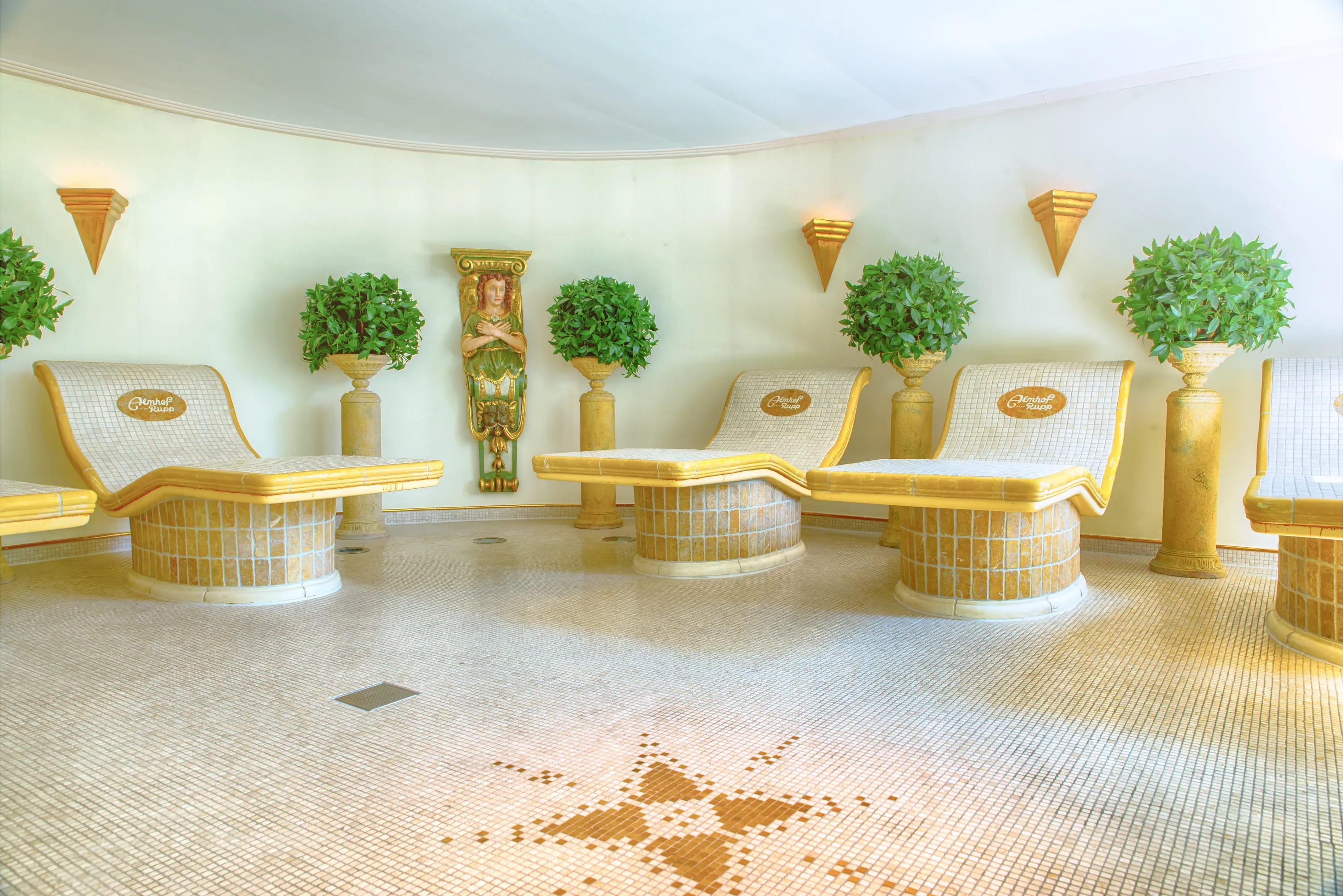 Relaxbereich mit goldenen Liegen um eine zentrale Statue, flankiert von Topfpflanzen und Wandlampen.