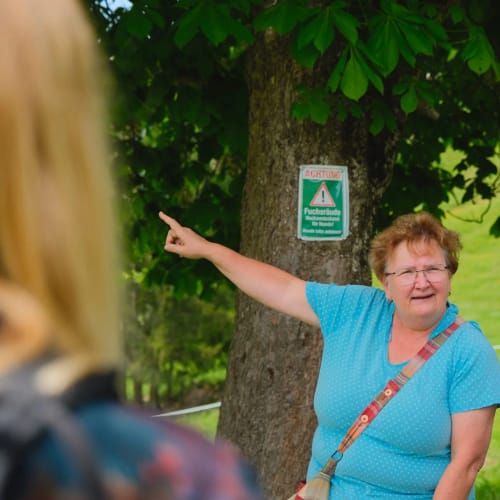 Frau in blauem Oberteil zeigt mit ausgestrecktem Arm neben einem Baum.