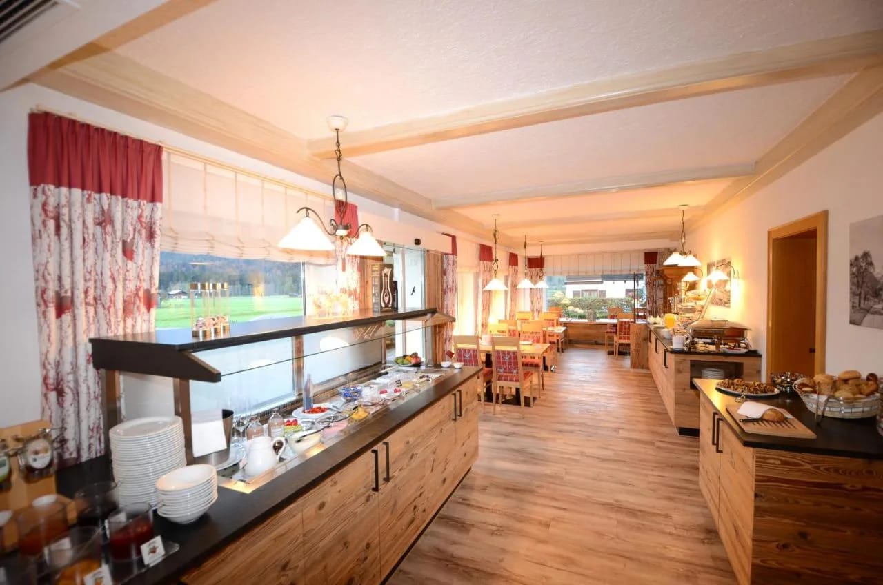 Heller Gastraum mit langer Frühstückstheke aus Holz, Lampen, Tischen und Blick nach draußen.