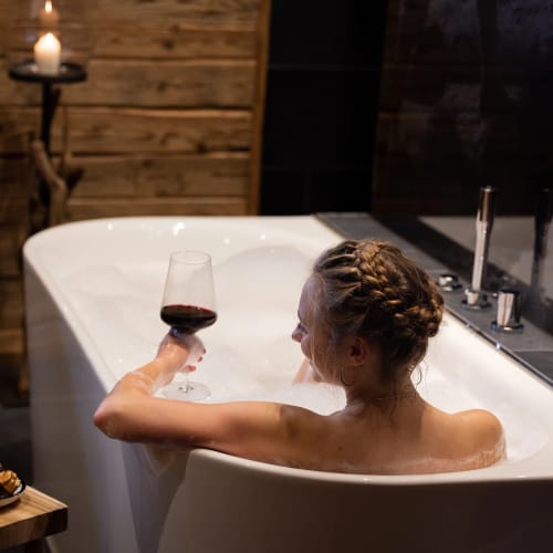 Person sitzt in Badewanne mit Glas Rotwein, Kerze brennt, Holzverkleidung im Hintergrund.