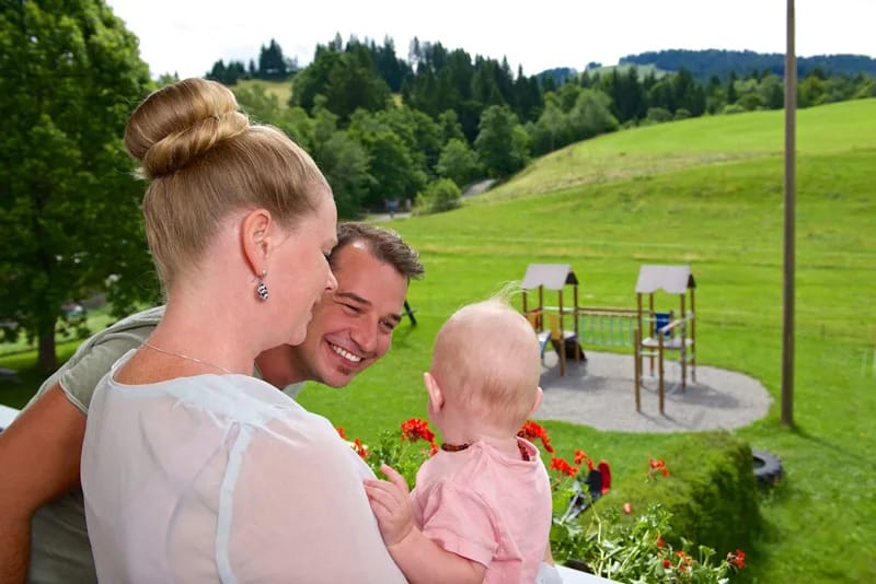 Familie mit Baby schaut lächelnd auf Spielplatz im Grünen