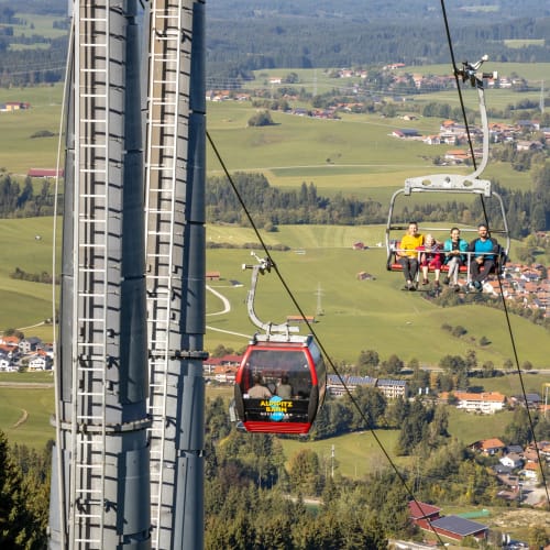 Personen in einer Seilbahnkabine über einer ländlichen Umgebung mit Feldern und Dorf im Hintergrund.
