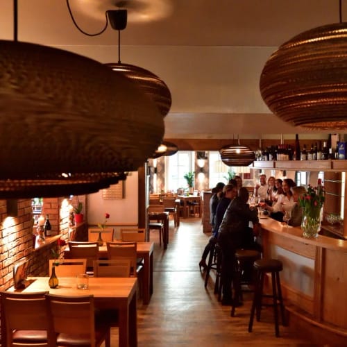 Innenansicht eines gemütlichen Restaurants mit Bar, Holztischen und großen Deckenspielders.