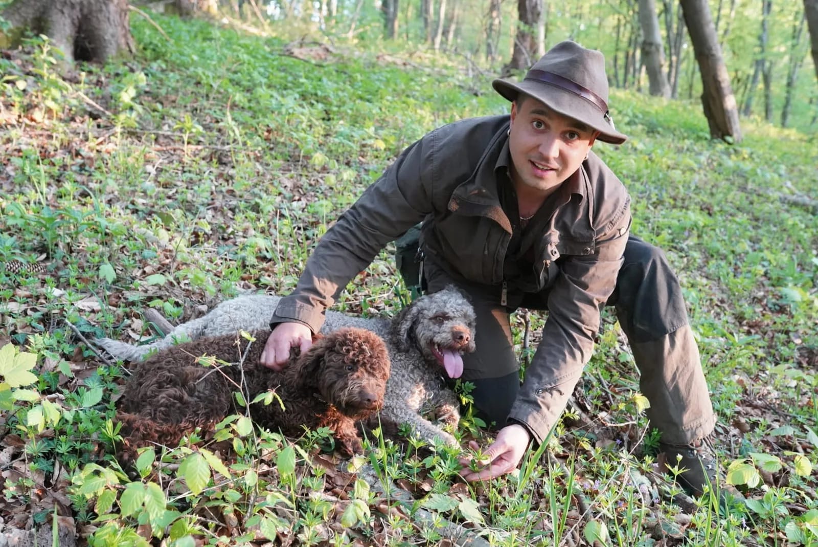 Person im Wald mit Hut kniet neben zwei kleinen Hunden auf Laub