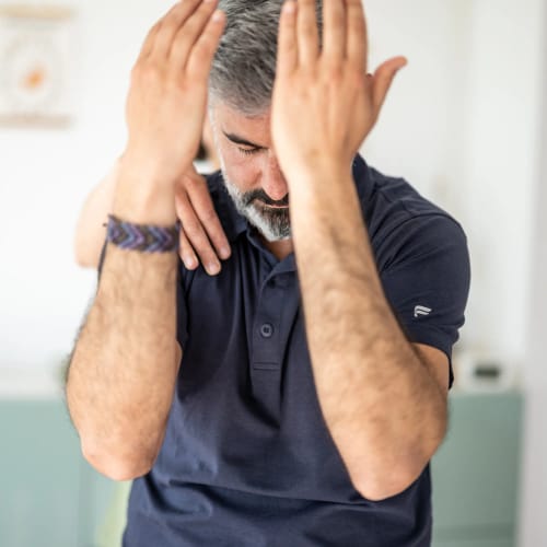 Mann mit grauem Bart greift sich den Kopf, wirkt gestresst in hellem Innenraum.