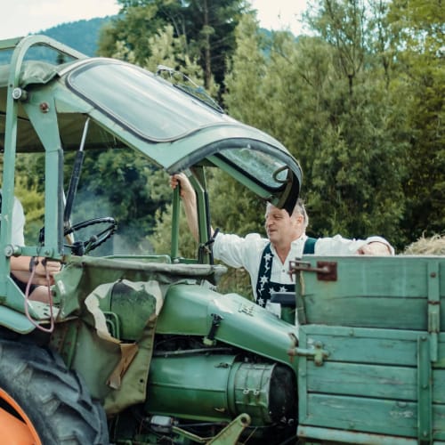 Zwei Männer in Trachten auf einem grünen Traktor mit Anhänger auf einer Wiese.