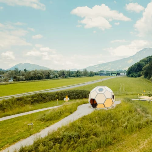 Geodome-Kuppel auf grüner Wiesenlandschaft mit Wegen und Bergen im Hintergrund.