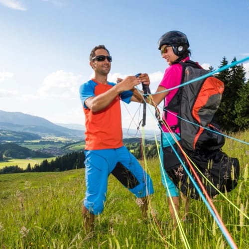 Zwei Personen mit Helmen und Ausrüstung bereiten Paragliding-Start auf einer Graswiese vor.