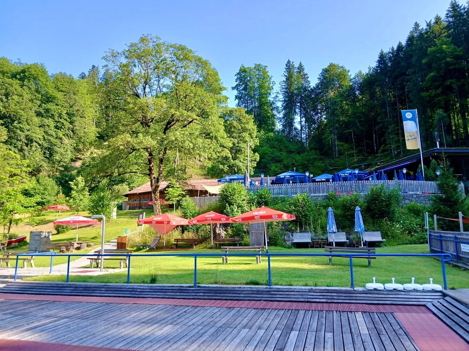 Sonniger Gartenteil mit Bäumen, roten und blauen Sonnenschirmen, Liegen und Holzdeck am Hang.
