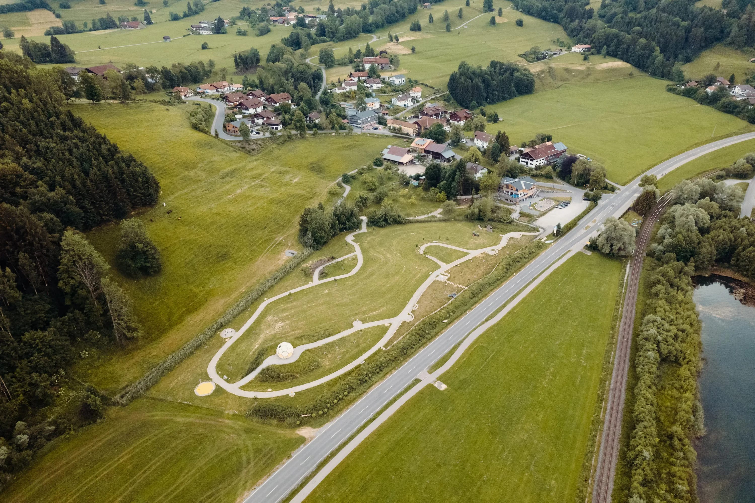Luftaufnahme eines Parkgeländes mit verschlungenen Wegen neben einer Hauptstraße, umgeben von Felder