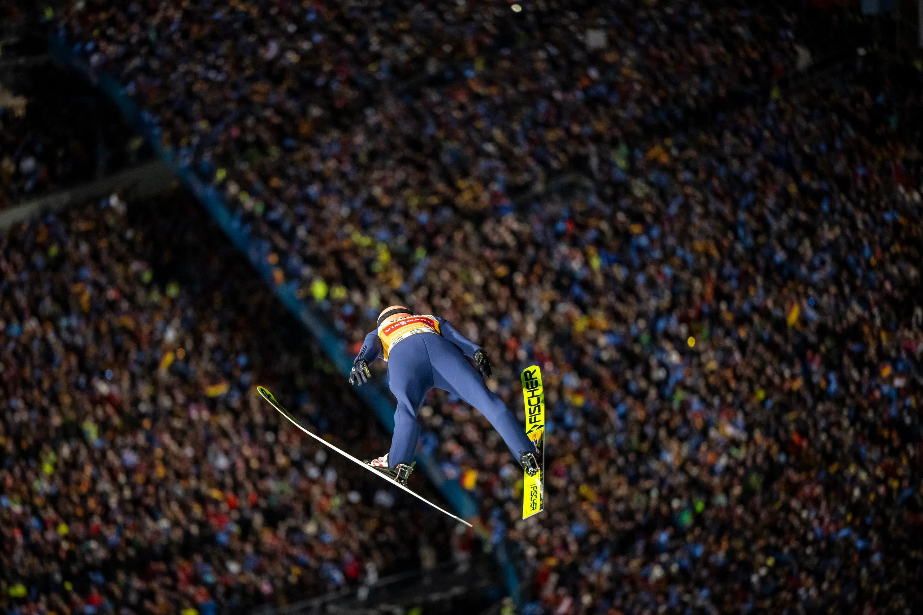 Skispringer in blauem Anzug fliegt im Sprung durch die Luft vor begeisterter Zuschauermenge im StadI