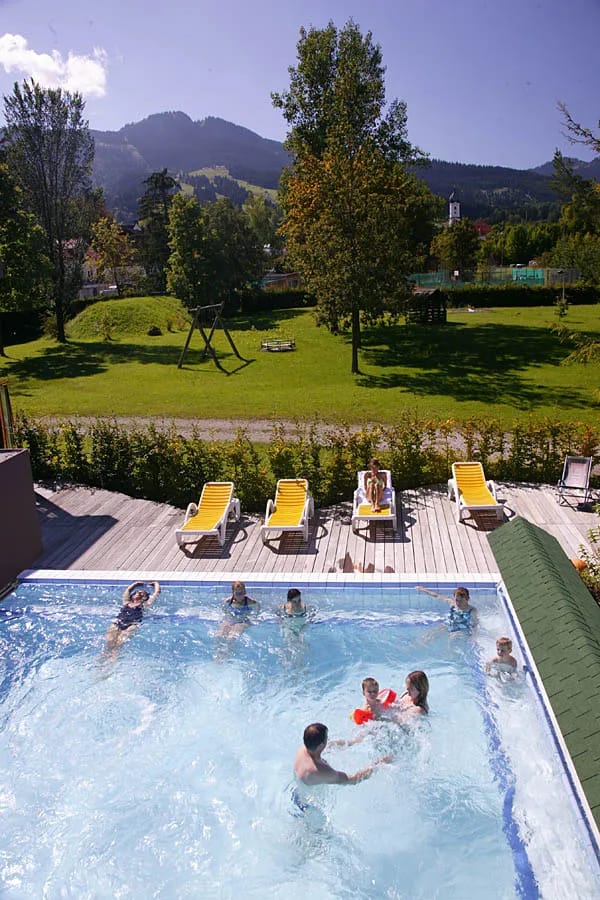 Außenpool mit mehreren Schwimmern, Liegen am Beckenrand und grünem Garten mit Bergen dahinter.