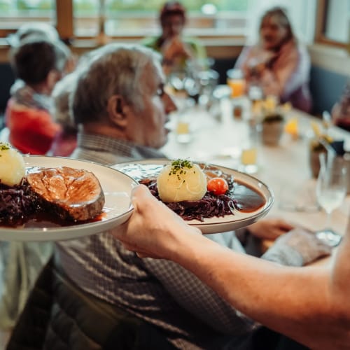 Kellner trägt zwei Teller mit Gericht zu einer Gruppe speisender Gäste in einem Restaurant.