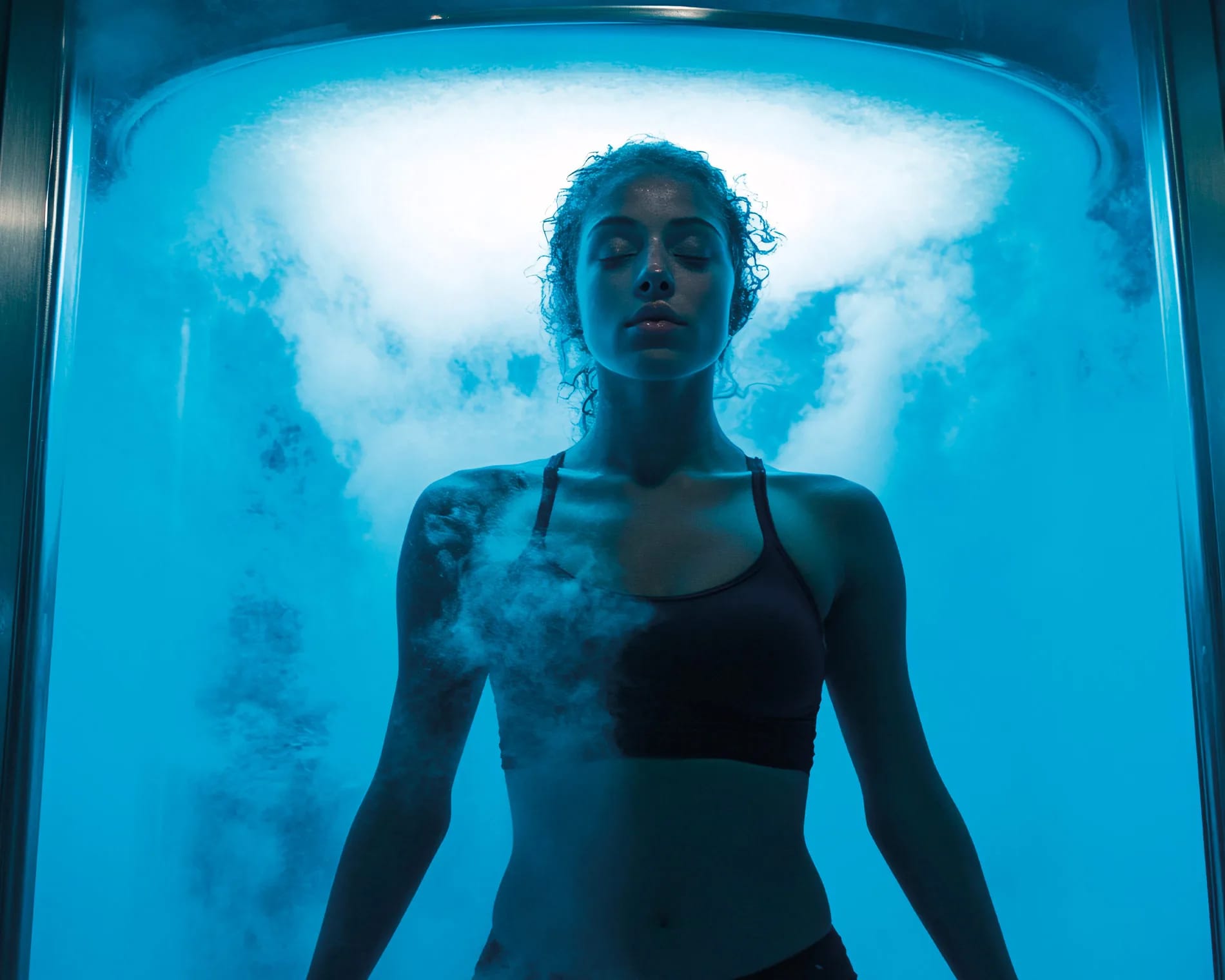 Junge Frau in Sport-BH in blauer Kryotherapie-Kammer, Nebel um sie herum.